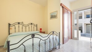 Une chambre à coucher avec un lit en métal, une table de chevet surmontée d’une lampe rouge et une porte donnant sur un balcon.