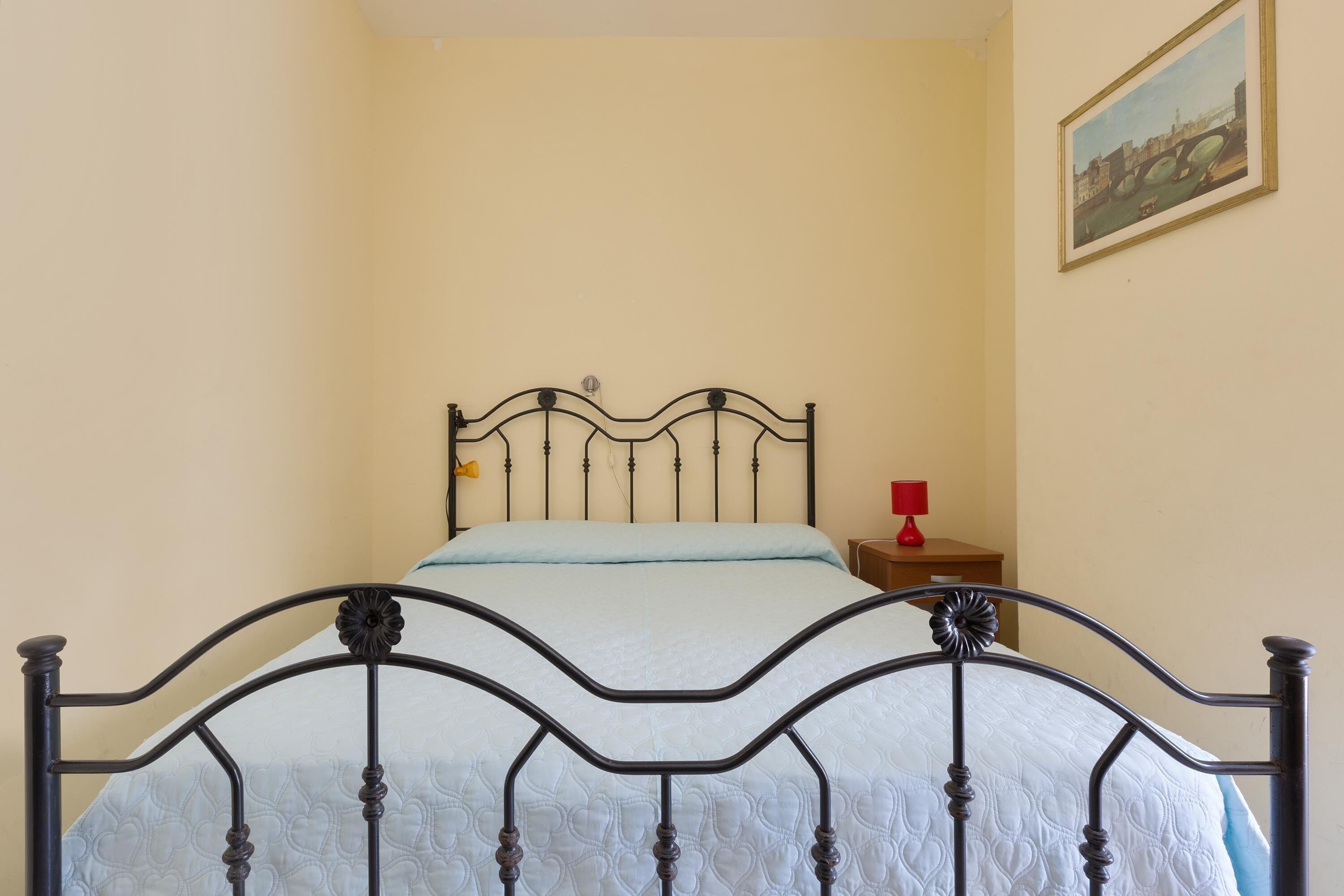 A bed with a metal frame and a quilted mattress in a room with plain walls and a single painting on the wall.
