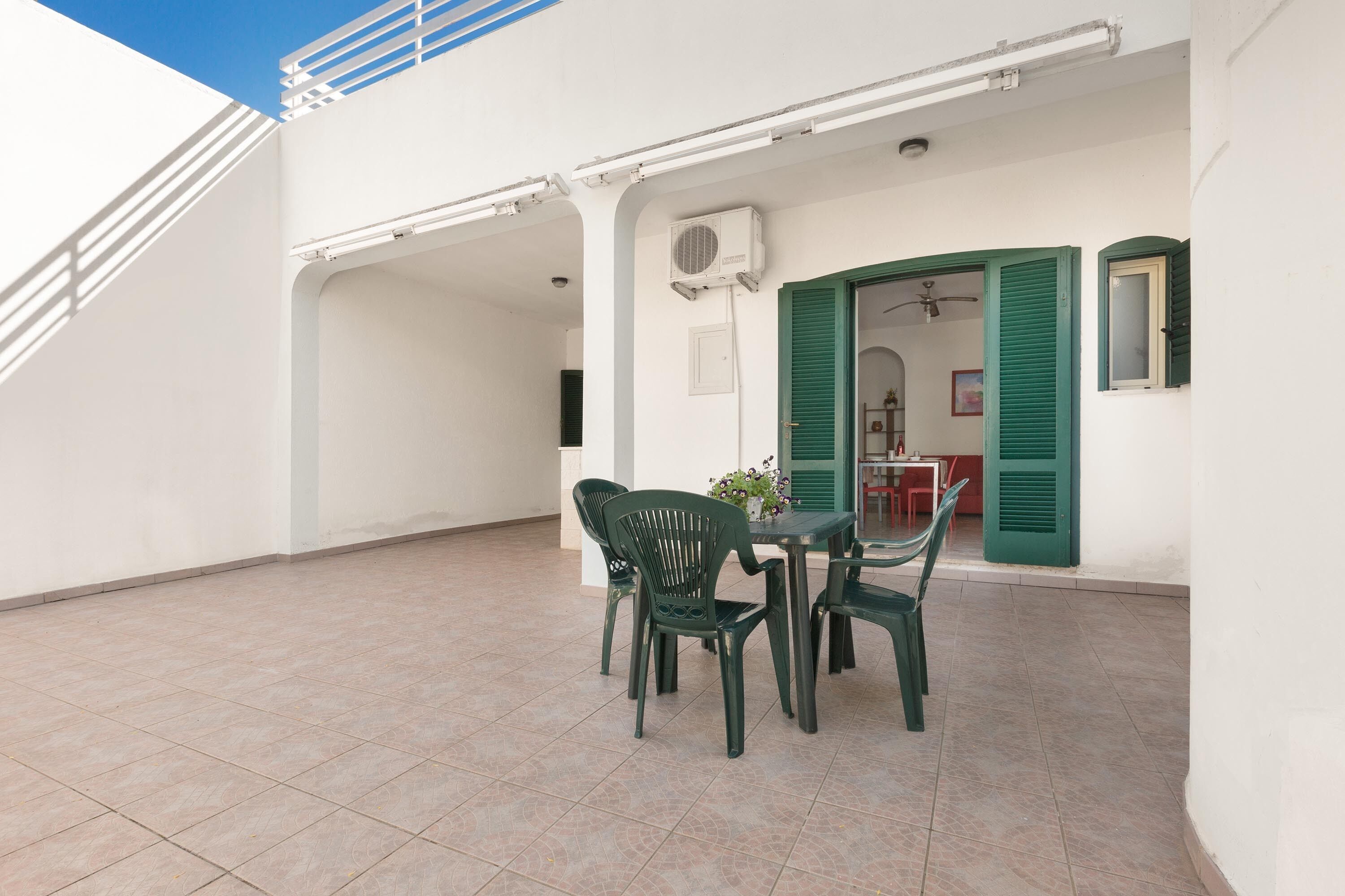 Um alpendre coberto com mesa e cadeiras, um aparelho de ar condicionado e vista para o edifício vizinho.