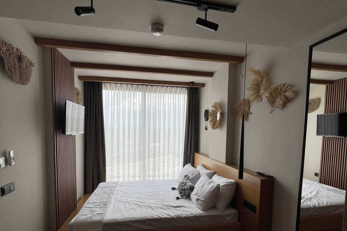 A modern hotel room with a large bed, a flat-screen TV, a ceiling-mounted light fixture, and a large mirror.