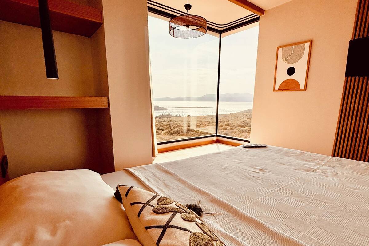 A bedroom with a large window, a bed with a patterned blanket, and a wall-mounted artwork.