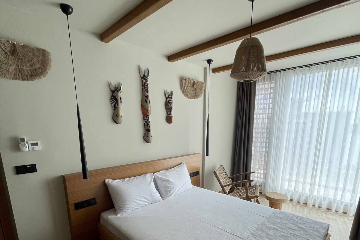 A bedroom with a wooden bed, white bedding, a ceiling fan, wall-mounted decor, and a window with sheer curtains.