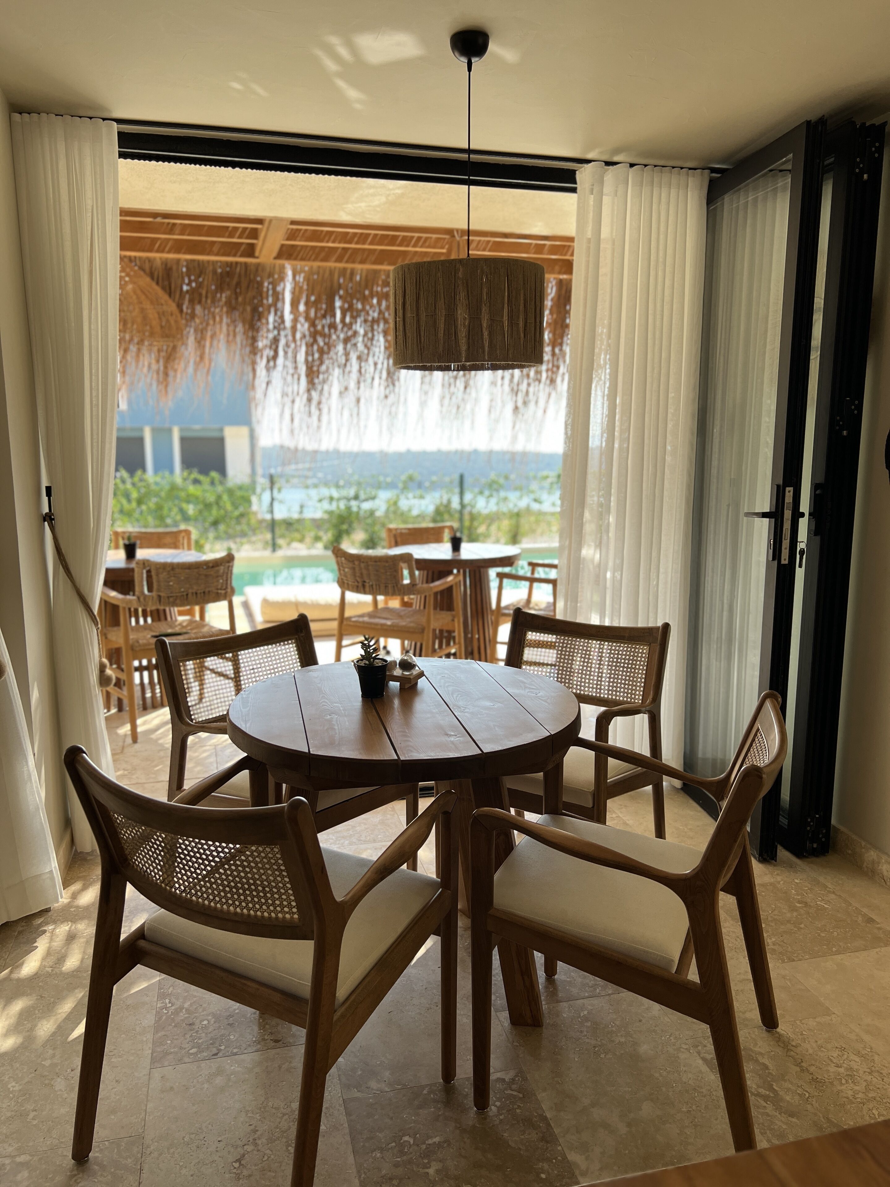 A dining area with a round wooden table and wicker chairs, overlooking a pool and greenery.
