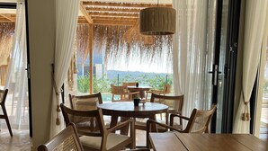 Uma sala de jantar com mesa e cadeiras de madeira, luminária pendente e vista para o verde lá fora.