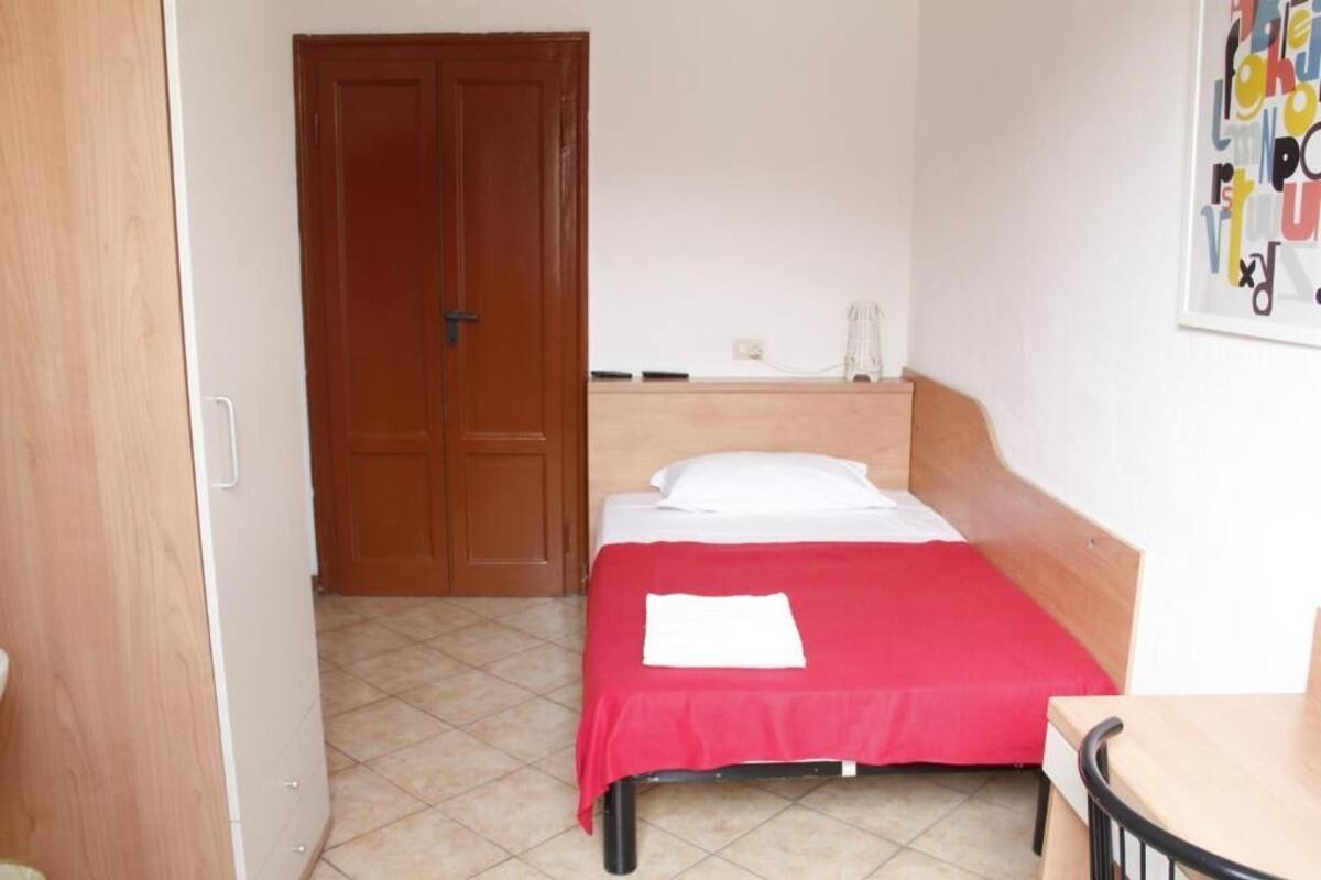 A bedroom with a bed, a wooden wardrobe, a desk, and a wall-mounted telephone.