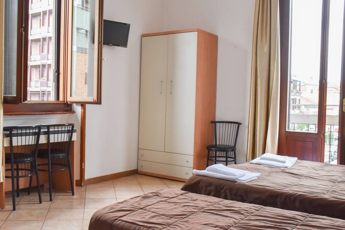 A bedroom with two beds, a desk with chairs, a wardrobe, and a TV on the wall.