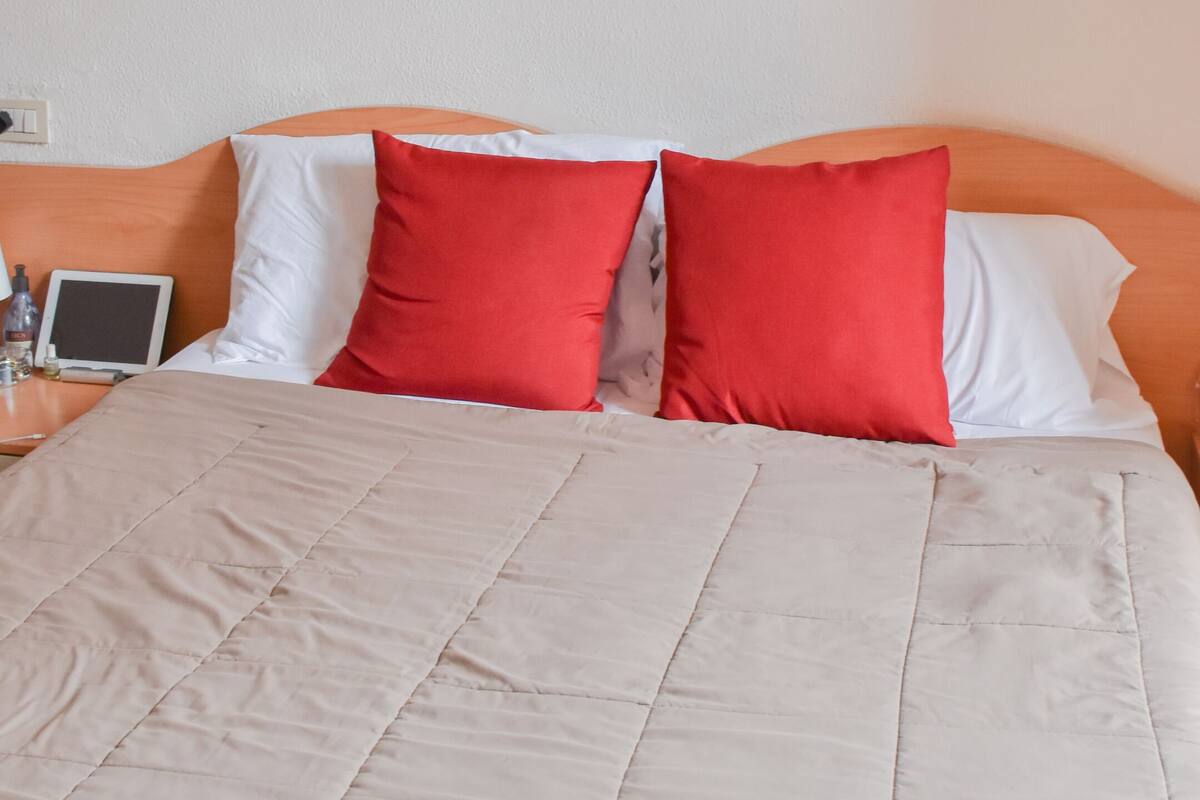 A hotel room with a large bed, two red pillows, a white pillow, a beige quilted bedspread, a wooden headboard, a bedside table with a glass, a tablet, and a framed picture on the wall.