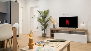A modern living room with a flat-screen TV, a dining table with books and a decorative item, a potted plant, and a minimalist kitchen in the background. - [7 min from airport] Design house with A/C and WiFi (ciampino)
