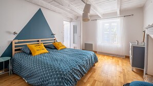Ein Schlafzimmer mit einem hölzernen Bett, einer blau-weißen Tagesdecke, zwei gelben Kissen, einer weißen Decke und einem Fenster mit Vorhängen.