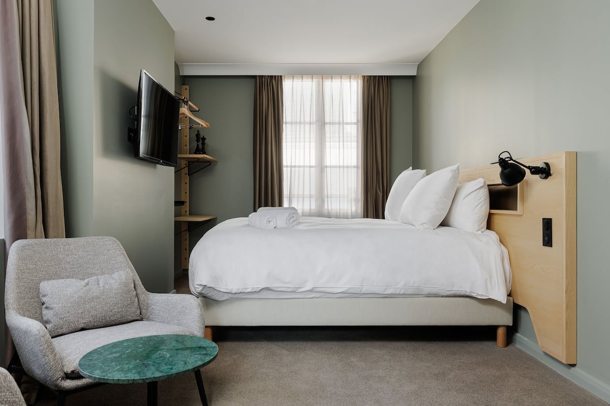 A hotel room with a bed, a chair, a TV mounted on the wall, and a window with curtains.