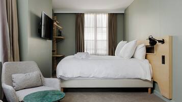A hotel room with a bed, a chair, a TV mounted on the wall, and a window with curtains.