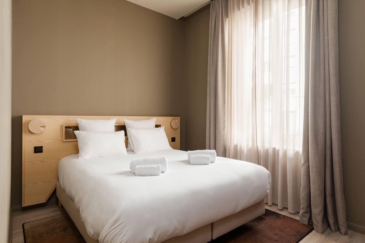 A neatly made bed with white linens and pillows, a wooden headboard, and a window with sheer curtains.