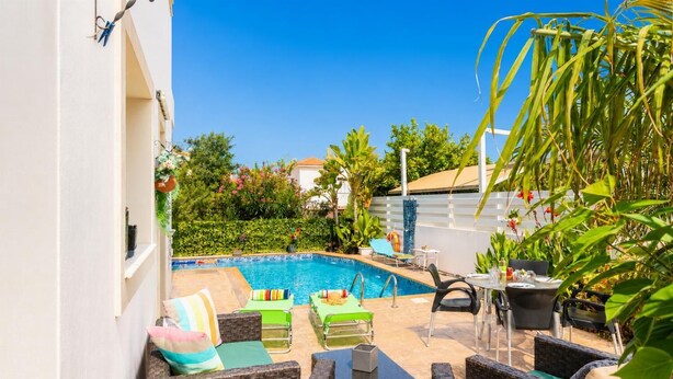 Ein Swimmingpool mit Liegestühlen und einem Tisch, umgeben von Grünpflanzen und einem weißen Gebäude.