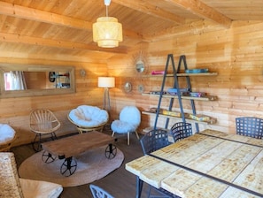 Acogedor interior de cabaña de madera con mesa de comedor de madera, sillas de mimbre, sillón redondo acolchado, lámpara de pie y estanterías con libros y objetos decorativos.