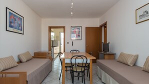 Una habitación pequeña y bien iluminada con una mesa y sillas de madera, dos camas individuales con ropa de cama color beige, un televisor sobre un mueble bajo y cuadros en las paredes.