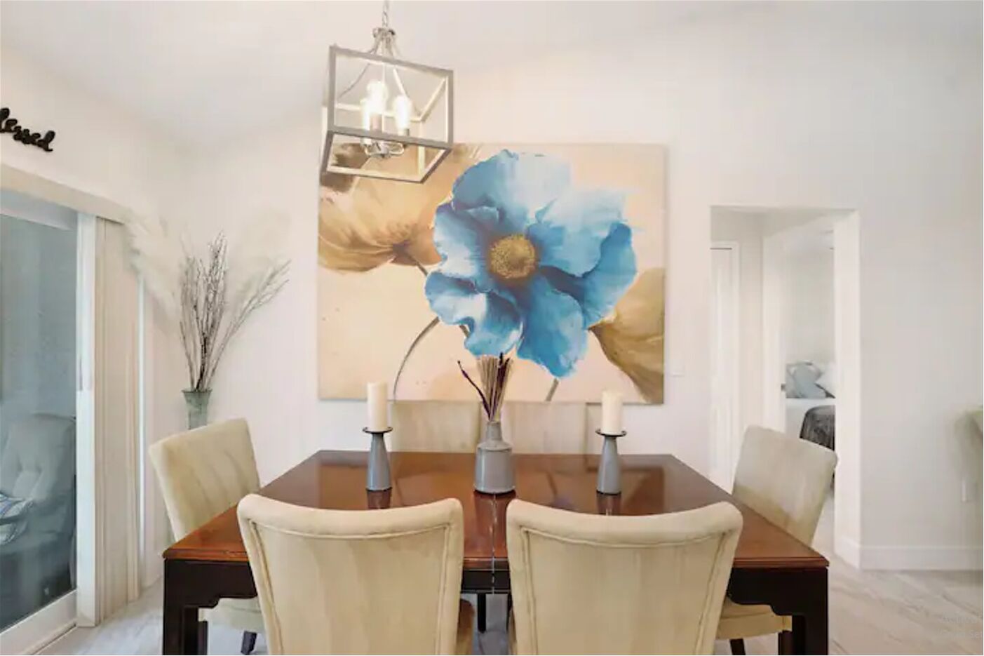 A dining room with a large floral painting, a chandelier, and a vase with candles on the table.