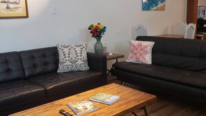 A living room with two black leather sofas, a wooden coffee table with books and a remote control, a vase with flowers, and a painting on the wall. - La Casita Solar (Clearwater)
