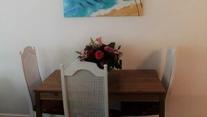 A dining area with a wooden table and white chairs, a wall-mounted painting of a beach scene, and a floral centerpiece on the table. - La Casita Solar (Clearwater)