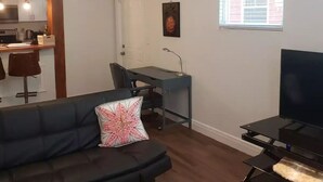 A modern living room with a black leather sofa, a desk with a computer, a television on a stand, a ceiling fan, and a window with a view of buildings. - La Casita Solar (Clearwater)