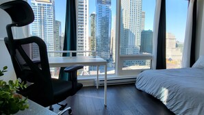 A modern hotel room with a large bed, a desk with a chair, and a view of tall buildings through large windows. - Lux Corner Unit-spa-gym-office-lounges (Montréal)