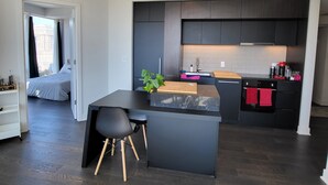 A modern kitchen with dark cabinetry, a central island, and a dining area with a black table and chairs. - Lux Corner Unit-spa-gym-office-lounges (Montréal)