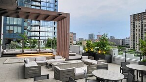 An outdoor seating area with round tables and chairs, a wooden pergola, and a view of a tall building with reflective windows. - Lux Corner Unit-spa-gym-office-lounges (Montréal)