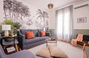 Ein Wohnzimmer mit grauem Sofa, einem kleinen runden Couchtisch, einer Lampe, einer Pflanze und einem tropischen Wandbild.