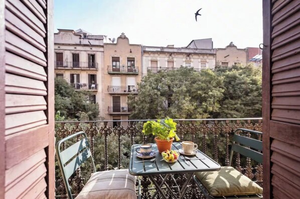 Un balcon avec une table dressée pour deux personnes, une plante en pot et un oiseau dans le ciel.