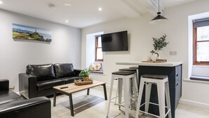 A modern living room with a black leather sofa, a wooden coffee table, and a TV mounted on the wall. There are two white bar stools and a small kitchen area with a sink and a potted plant. - Llety Pen Deitsh 2 (Caernarfon)