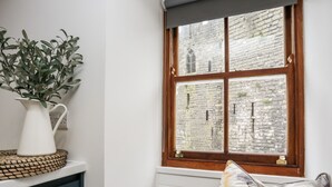A corner room with a window overlooking a stone building, a white vase with green leaves, and a bench with a floral pillow. - Llety Pen Deitsh 2 (Caernarfon)