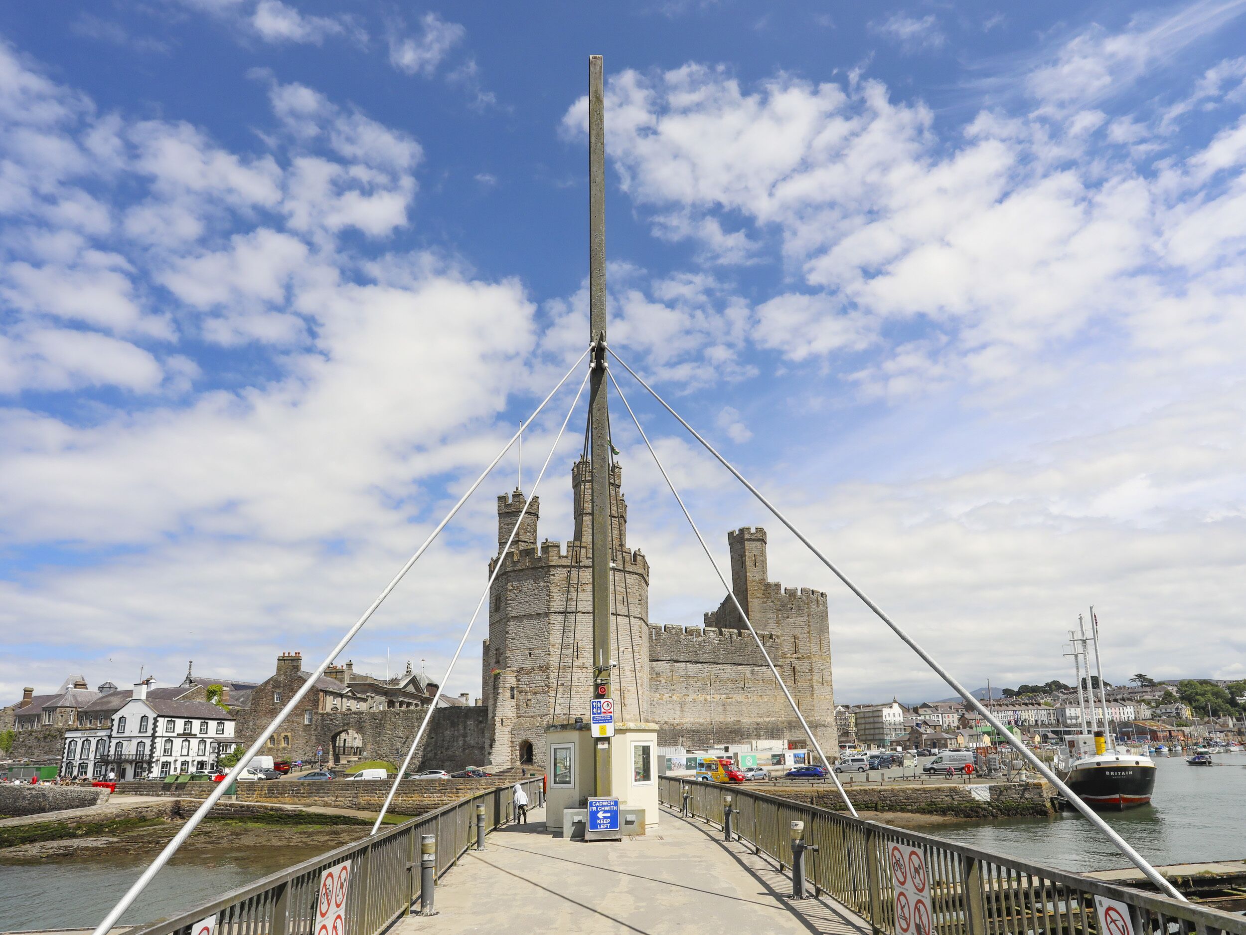 Un puente con una torre central alta, un edificio de piedra al fondo y un puerto deportivo con barcos.