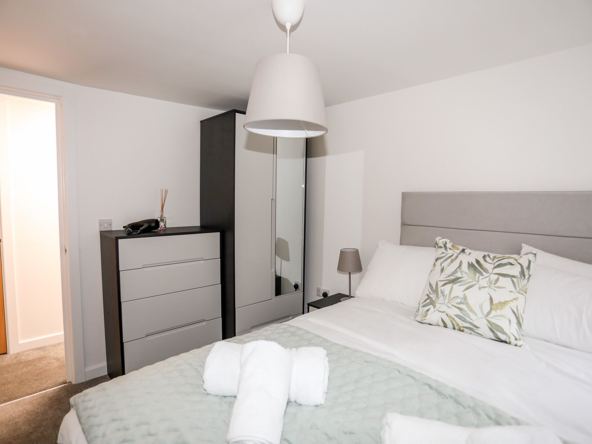 Une chambre d’hôtel bien rangée, avec un lit, une commode, un miroir et une table de chevet avec une lampe.