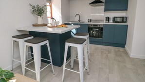 A modern kitchen with a central island, bar stools, a sink, and built-in appliances. - Llety Pen Deitsh 2 (Caernarfon)