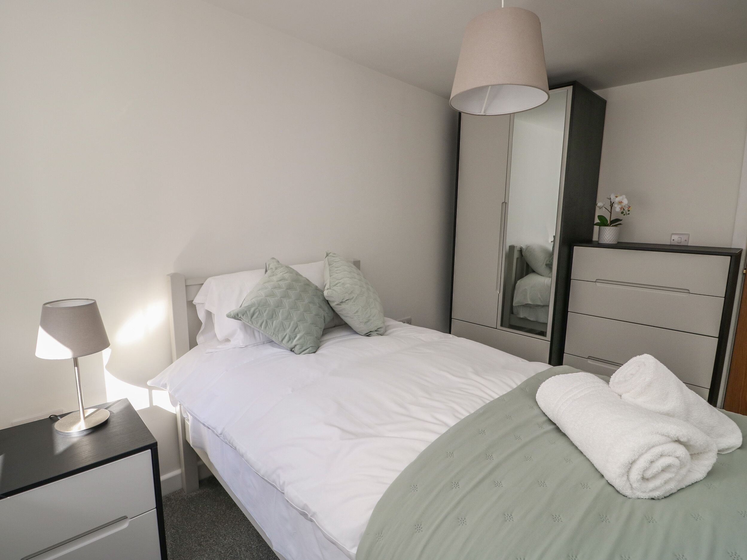 Un lit bien fait, avec des oreillers, une lampe de chevet, une commode avec des tiroirs, un miroir et une serviette posée sur le lit.