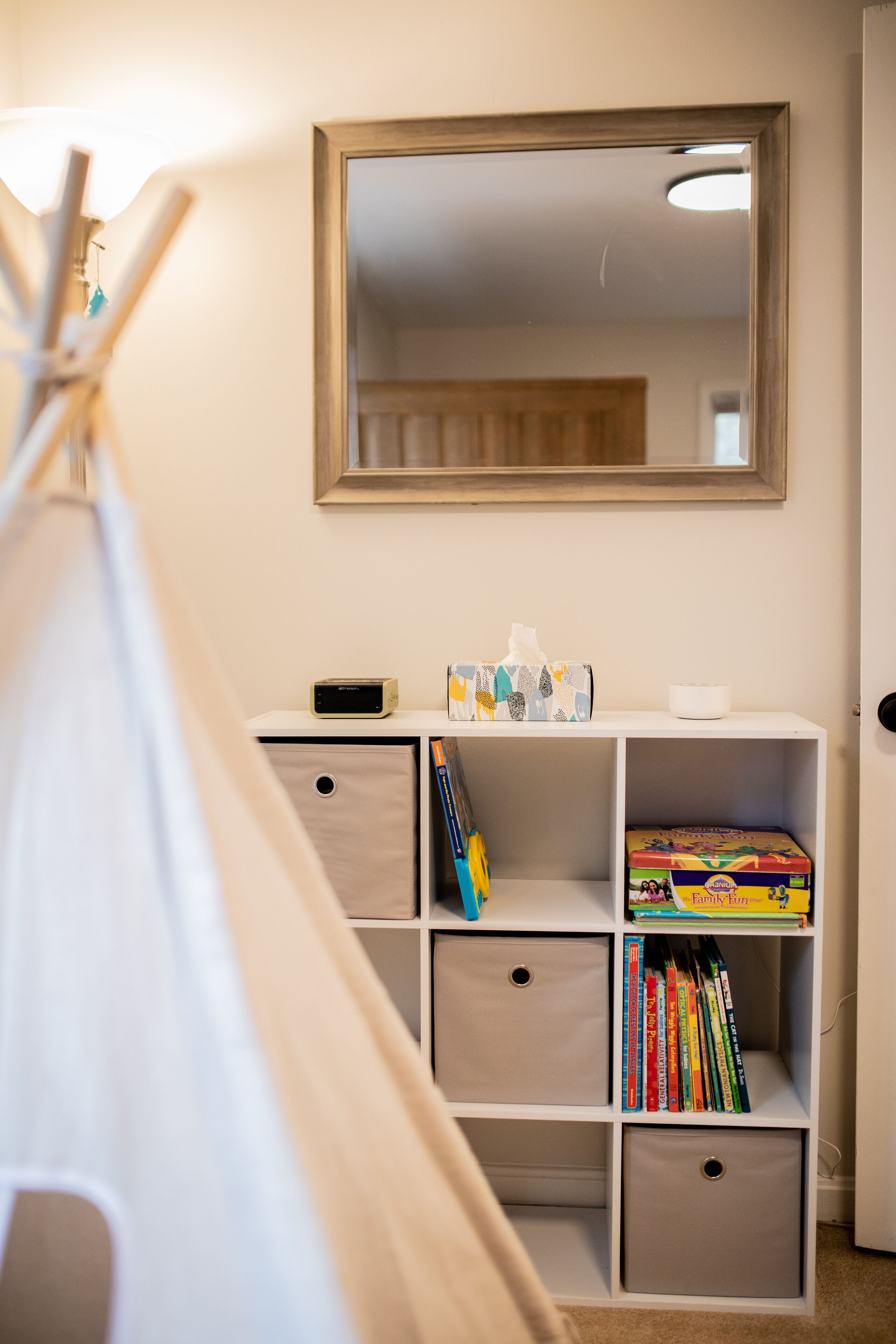A room with a white cabinet containing books and a box, a mirror on the wall, and a tent in the foreground.