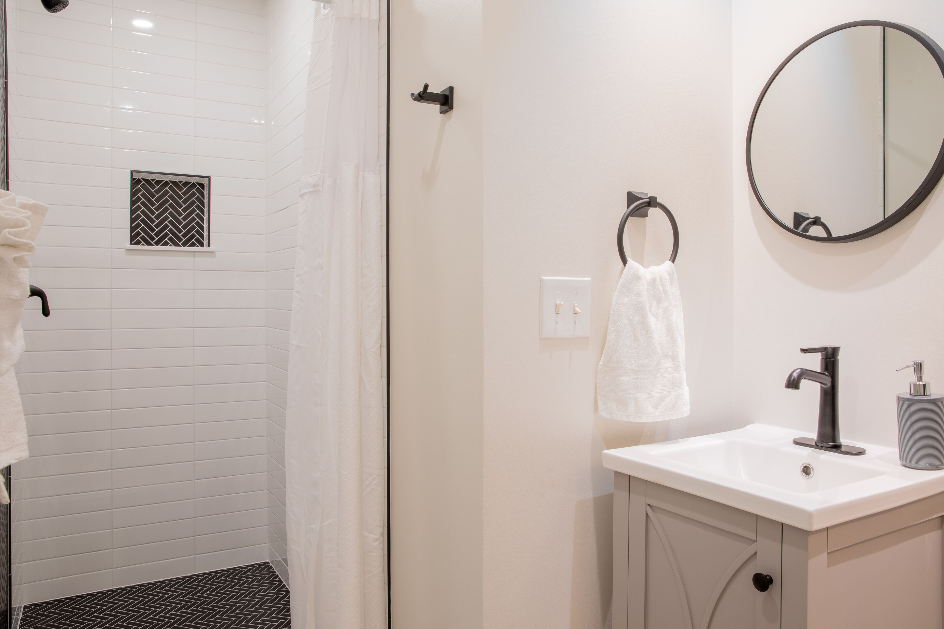 A modern bathroom with white tiled walls, a shower with a black and white pattern, a white sink with a black faucet, and a round mirror.