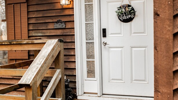 Une cabane en bois avec une porte d’entrée blanche, une pancarte de bienvenue et un escalier en bois menant à l’entrée.