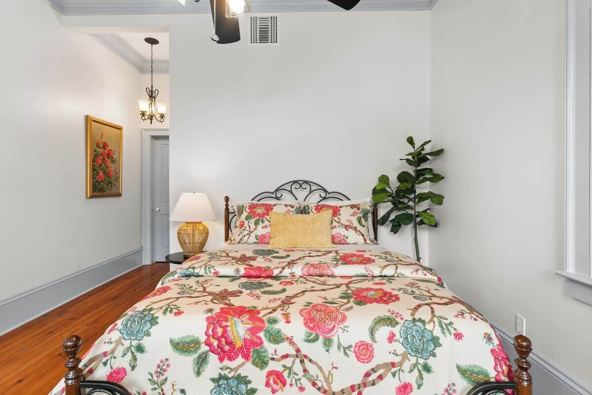 A bedroom with a floral bedspread, a nightstand with a lamp, a wall-mounted picture, and a potted plant.