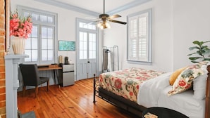 Quarto com piso de madeira, cama com roupa de cama floral, escrivaninha com cadeira, ventilador de teto e janela com persianas.