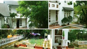 Un edificio blanco con tejado de baldosas, rodeado de abundante vegetación y una cerca de piquetes blancos.