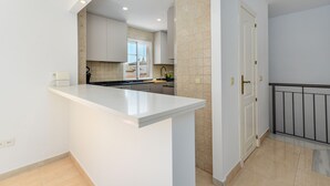 A modern kitchen with white cabinetry, a central island, and a view into the dining area. - Duquesa Regent Penthouse 2435 Sea-view Duplex (Manilva)