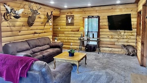 Ein Holzhaus-Interieur mit einem braunen Ledersofa, einem Couchtisch mit Pflanze, einem aufgehängten Hirschkopf, einem aufgehängten Vogel und einem Flachbildfernseher an der Wand.