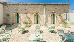 Un patio con suelo de piedra, mobiliario de exterior verde y un edificio de piedra con puertas arqueadas.