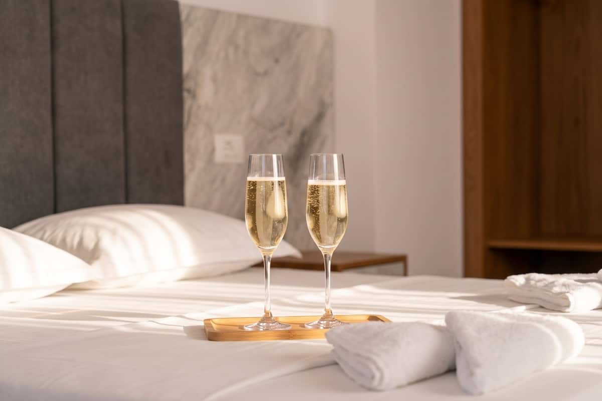 Two champagne glasses on a tray on a bed with white linens, a wooden headboard, and a wooden wardrobe in the background.