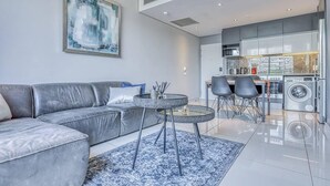 A modern living room with a grey sofa, a coffee table, and a rug. There is a kitchen area with a washing machine and a dining table with chairs. - Luxury 1 Bed Apartment at The Capital on the Park (Johannesburg)