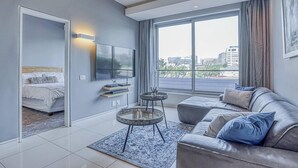 A modern living room with a grey sofa, a flat-screen TV, a coffee table, and a bed with a headboard. - Luxury 1 Bed Apartment at The Capital on the Park (Johannesburg)