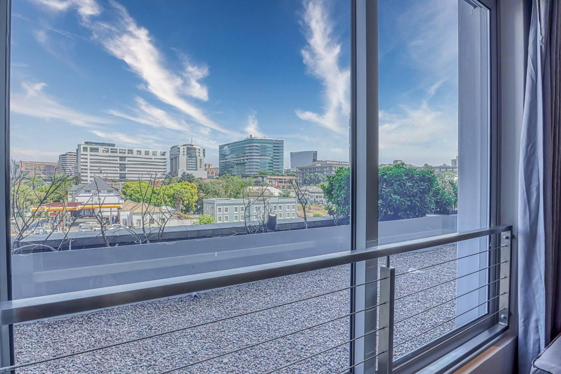 A cityscape view from a window with a metal railing, featuring modern buildings and a clear sky.