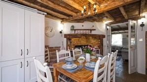 Ein Essbereich mit einem Holztisch, gedeckt für vier Personen, umgeben von weißen Stühlen. Ein Backsteinofen mit Sims ist sichtbar, und eine Uhr hängt an der Wand.