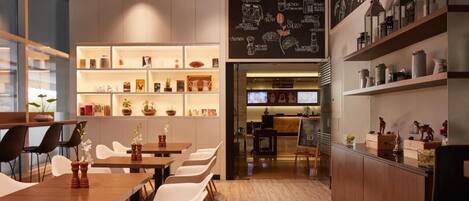 A modern cafe with wooden tables and white chairs, shelves with various items, and a chalkboard menu.