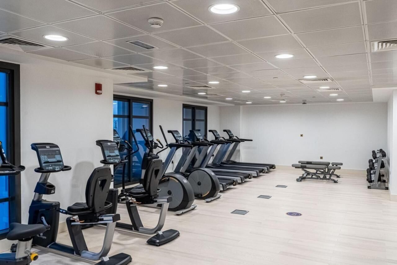 Ein Fitnessraum mit mehreren Laufbändern und Ergometer, einem Holzboden und großen Fenstern.